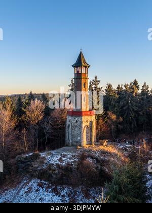 Der Aussichtsturm Stepanka steht hoch im Isergebirge. Das Sonnenuntergangslicht wirft Schatten auf den schneebedeckten Boden. Bäume umgeben den Turm und schaffen eine wunderschöne Landschaft. Stockfoto