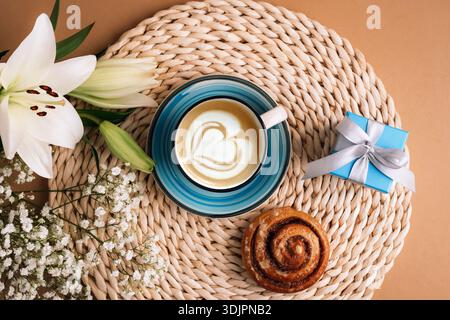 Tasse Kaffee mit Zimtbrötchen, Blumen und kleiner Geschenkbox auf gewebtem Tischset. Happy Valentines Day-Konzept. Draufsicht, flach. Stockfoto