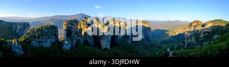 Panoramablick auf die Meteora-Klöster und Felsformationen, die sich über einem bewaldeten Tal mit fernen Bergen unter einem blauen Himmel erheben, Thessalien, Griechenland Stockfoto