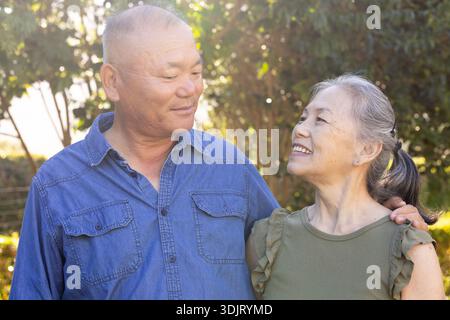 Seniorenpaar steht in der Nähe, umarmen und lächeln im sonnigen Garten mit Denim-Hemd und olivfarbenem Oberteil Stockfoto