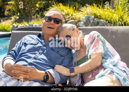 Ein älteres asiatisches Paar liegt in der grauen Lounge am Pool, lächelt und trägt eine Sonnenbrille Stockfoto
