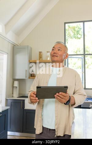 Ein älterer asiatischer Mann mit Tablette, der auf der Kücheninsel in der Nähe des Fensters steht, mit beigem Overshirt Stockfoto