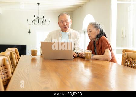 Älteres asiatisches Paar sitzt zu Hause an einem langen Holztisch mit Laptop und Glasbecher in der Nähe Stockfoto