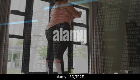 Stehende Frau auf dem Hocker zum Fenster zu Hause, mit rosa Sportoberteil und Leggings, Code-Overlay Stockfoto