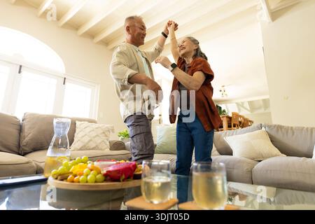 Verschiedene Seniorenpaare tanzen im Wohnzimmer in lässigen Klamotten neben einem Glastisch mit Obst. Sonnenlicht, Bogen, Fenster, Balken, offener Plan, Sofa, c Stockfoto