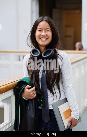 Porträt einer lächelnden jungen Frau mit schnurlosen Kopfhörern um den Hals, die im Universitätskorridor steht Stockfoto