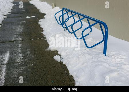 An einem klaren Wintertag steht ein Fahrradträger auf einem schneebedeckten Gehweg. Fußabdrücke sind auf dem Pfad sichtbar. Stockfoto