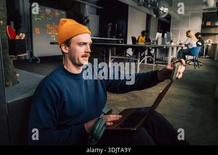 Männlicher Geschäftsunternehmer, der Selfie über das Smartphone macht, während er mit Laptop im Technikbüro sitzt Stockfoto