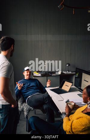 Glücklicher Geschäftsexperte, der mit Kollegen am Schreibtisch im Technikbüro diskutiert Stockfoto