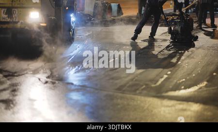 Arbeiter, der nachts frischen Asphalt auf der Stadtstraße verdichtet, Dampf steigt an Stockfoto