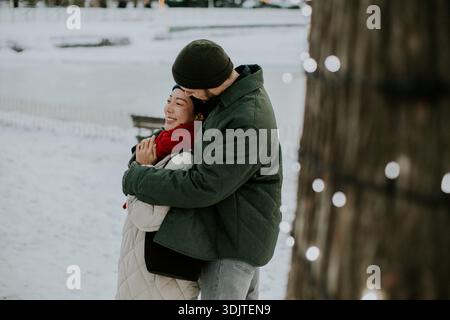 Eine Koreanerin und ein Kaukasier stehen nahe in einem verschneiten Park. Sie teilen einen Moment der Freude, während sie umarmen und die Winteratmosphäre und den Urlaub genießen Stockfoto