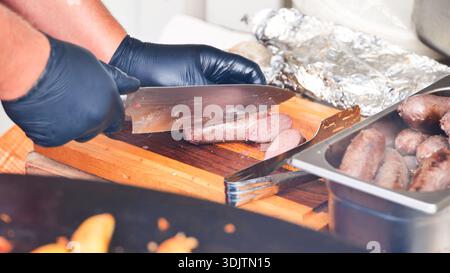 Die Hände des Küchenchefs tragen schwarze Handschuhe und schneiden gegrillte Würstchen auf einem Holzbrett Stockfoto