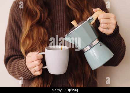 Frau, die heissen Kaffee aus der Mokakanne in eine Keramikbecher zu Hause gießt. Gemütliche Morgenroutine mit frisch gebrühtem Espresso. Zubereitung eines warmen Getränks. Stockfoto