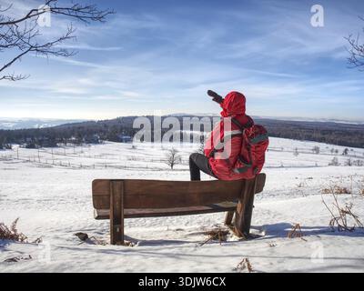 Seitenansicht des Mannes, der am Wintertag im Schnee spaziert, Berglandschaft. Wanderer in Rot mit rotem Rucksack. Winterliches Wanderreisekonzept Stockfoto