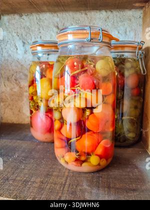 Gläser mit verschiedenen eingelegten Tomaten und Gemüse werden in einer rustikalen Küche auf einem Holzregal gegen eine strukturierte Steinwand gestellt Stockfoto
