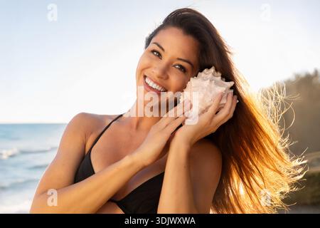 Ein wunderschönes, braunes, multiethnisches Modell mit gemischter Rasse genießt das Rauschen des Ozeans, während es an einem tropischen Strand eine Muschel ans Ohr hält Stockfoto