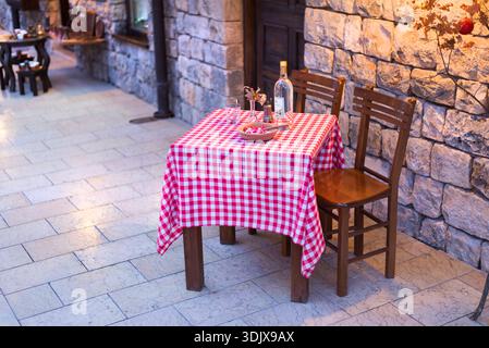 Gemütlicher Restauranttisch im Freien mit roter karierter Tischdecke, Holzstühlen, Weinflasche, Gläsern und warmer Abendatmosphäre in traditioneller Steinstraße Stockfoto