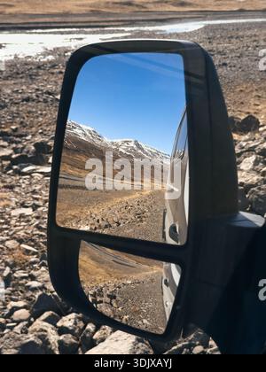 Blick durch den Autospiegel einer Straße durch die karge Landschaft Islands mit schneebedeckten Bergen im Hintergrund Stockfoto