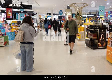 Reisende mit Rollgepäck spazieren durch Auckland Duty Free innerhalb des Flughafens Auckland, Neuseeland, vorbei an Einzelhandelsanzeigen und Schildern. Stockfoto