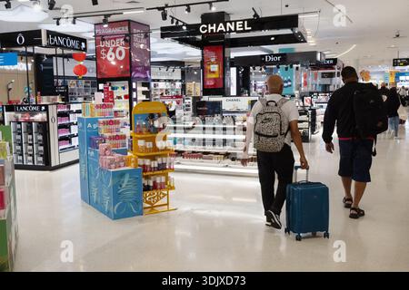 Reisende mit Rollgepäck spazieren durch Auckland Duty Free innerhalb des Flughafens Auckland, Neuseeland, vorbei an Einzelhandelsanzeigen und Schildern. Stockfoto