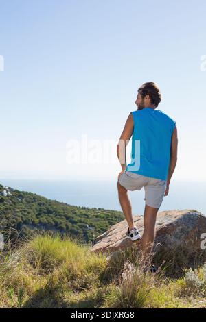 Der Mensch steht mit einem Fuß auf dem Felsen und blickt auf das ruhige blaue Meer vom Küstenhang. Wanderer, Landschaft, Küste, Aussichtspunkt, Natur, Einsamkeit, Sonnenschein Stockfoto