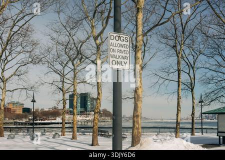 Ein Schild weist darauf hin, dass Hunde auf dem gepflasterten Weg in einem Winterpark in der Nähe des Wassers mit Bäumen herumlaufen können. Stockfoto
