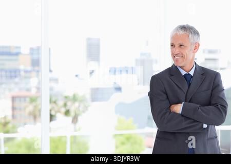 Reifer Mann, der neben dem raumhohen Fenster in einem modernen Büro steht, Anzug und Krawatte trägt, Kopierraum. Executive, Stadtlandschaft, Tageslicht, Highkey, Balkon, Stockfoto