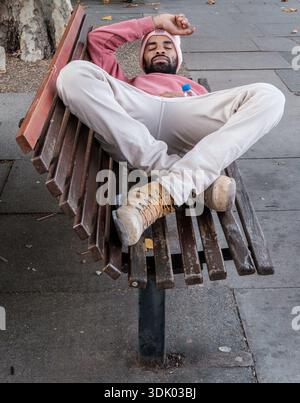 Entspannter bärtiger Mann in rosafarbenem Hoodie und beiger Jogginghose lässt sich im Sommer bequem auf einer Holzbank im Freien ausstrecken. Stockfoto
