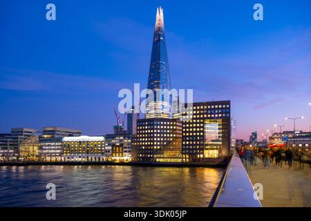 Der Shard erhebt sich über der Themse, wenn der Tag in London zur Nacht wird. Die Menschen bewegen sich entlang des Gehwegs und genießen die Stadt, während die Lichter zu leuchten beginnen. Diese Szene fängt das urbane Leben ein. Stockfoto
