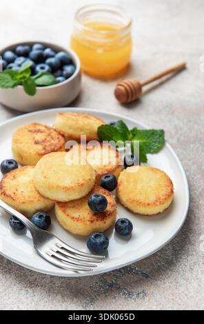 Käsepfannkuchen oder Syrniki, mit Heidelbeeren und Minze auf einem weißen Teller mit Honigtopf Stockfoto