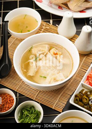 Appetitliche Schüssel Wontons in leichter Brühe, begleitet von verschiedenen Gewürzen und Gewürzen, serviert auf einer Bambusmatte. Stockfoto