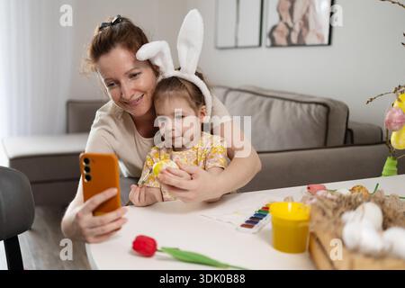Lächelnde Mutter und Tochter bereiten sich zuhause freudig auf Ostern vor. Das Mädchen trägt Hasenohren und hält ein bemaltes Ei, während ihre Mutter ihre festliche Aktivität mit einem Mobiltelefon festnimmt. Stockfoto