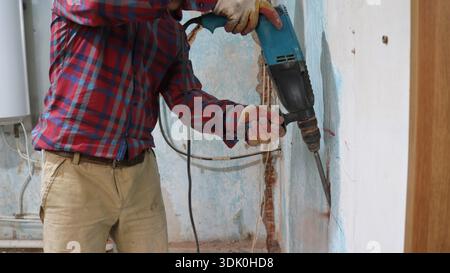 Ein Mann in brauner Hose und rotem kariertem Hemd hält einen Bohrer und meißt mit viel Mühe eine lange vertikale Nut in der Wand, um Drähte zu verstecken Stockfoto