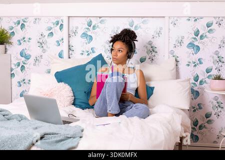 Eine junge Frau sitzt bequem auf ihrem Bett und konzentriert sich voll auf ihr Notebook, während sie Kopfhörer trägt. Der ruhige Raum hat wunderschöne Pflanzen und weiche Texten Stockfoto