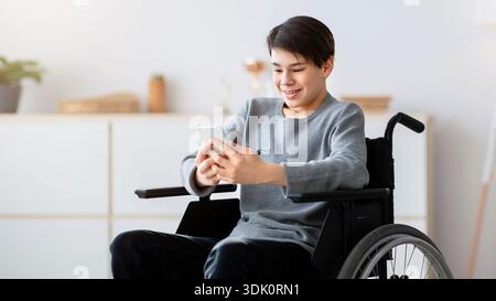Junger asiatischer Mann mit Smartphone, während er drinnen im Rollstuhl sitzt Stockfoto