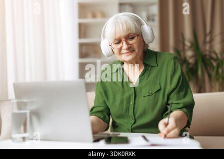 Ältere Frau, die Kopfhörer trägt, während sie zu Hause einen Laptop benutzt Stockfoto