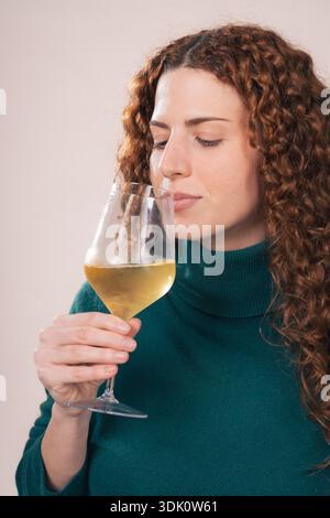 Eine junge Frau mit lockigen roten Haaren, die einen grünen Rollkragen trägt, riecht ein Glas Weißwein in einem Atelierporträt, das die Weinprobe und hervorhebt Stockfoto
