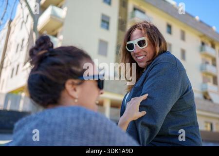 Lächelnde Frau, die weiße Sonnenbrille trägt, mit einem Freund im Freien in einer urbanen Umgebung spricht und lacht, einen glücklichen Moment teilt Stockfoto