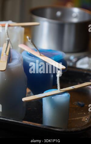 Frisch gegossene handgemachte Kerzen in verschiedenen Blau- und Weißtönen kühlen in den Formen ab, mit Holzstäben, die die Dochte an Ort und Stelle halten. Stockfoto