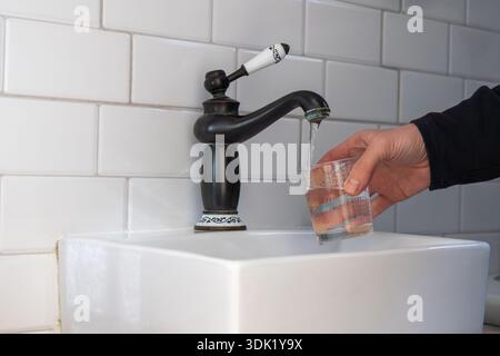 Eine Person, die ein klares Glas mit Trinkwasser aus einem schwarzen Wasserhahn im Vintage-Stil über einem weißen Waschbecken vor der Wand mit weißen Fliesen füllt. Reinheitsbegriff und Stockfoto