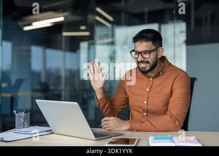 Junger Geschäftsmann, der von seinem modernen Büro aus an einer Videokonferenz teilnimmt, lächelt und auf einem Laptop Kollegen oder Kunden zuwinkt und dabei die Aufmerksamkeit von Stockfoto