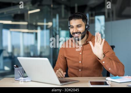 Selbstbewusster bärtiger Mann, der ein Headset trägt und lächelt, an einem Schreibtisch in einem modernen Büro sitzt und aktiv an einem erfolgreichen virtuellen Bus teilnimmt Stockfoto