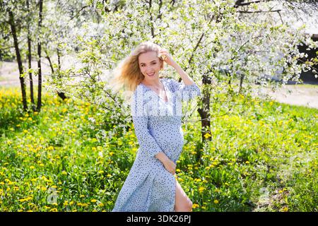 Eine schöne junge schwangere Frau in einem Kleid mit Strohhut geht durch den Frühlingsgarten. Glückliche Schwangerschaft. || Modell freigegeben Stockfoto