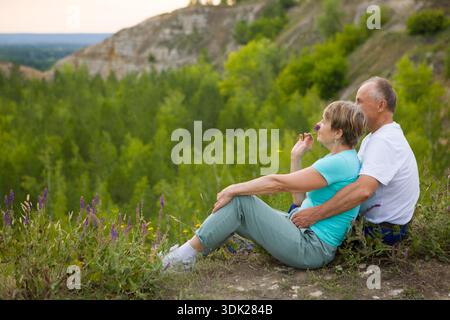 Ein älteres Ehepaar, das auf einem Berg sitzt, bewundert die Landschaft bei Sonnenuntergang. Seniorenpaar spazieren in der Natur. Reisetourismuskonzept. Verbringt im Sommer Zeit in der Natur. || Modell freigegeben Stockfoto