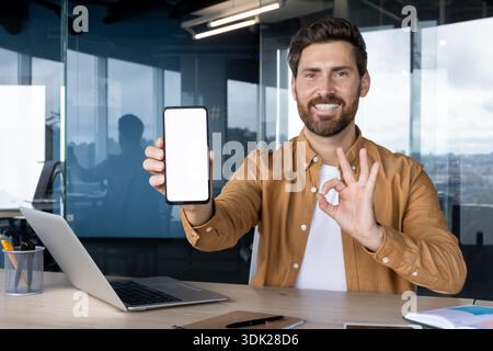 Lächelnder professioneller Mann, der ein Mobiltelefon mit einem weißen Display für App- oder Website-Mockup präsentiert, während er in seinem Büro sitzt Stockfoto