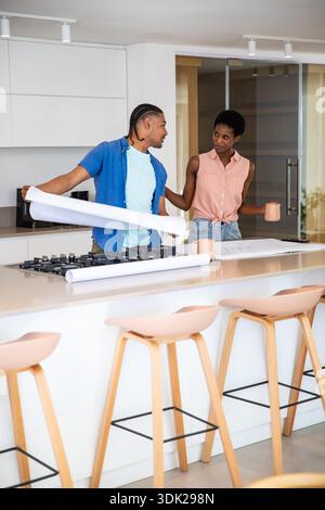 Verschiedene Paare, die Blaupausen auf der Kücheninsel in der Nähe des Gasherds lesen, während sie Keramikbecher halten. Architekturdesign, Hausrenovierung, modernes Interieur, Zusammenarbeit, Planung, Lifestyle, Renovierungskonzept || Modell veröffentlicht Stockfoto