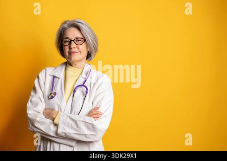 Ärztin, die mit gekreuzten Armen steht, Laborkittel und Stethoskop auf gelbem Hintergrund trägt Stockfoto