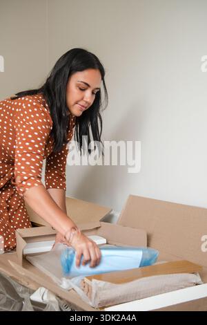 Eine Frau packt eine Kiste aus und steckt eine blaue Plastiktüte hinein. Sie trägt ein braunes Kleid mit Punktmuster Stockfoto