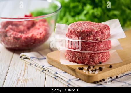 Stapel roher Rindfleischburgerpasteten, getrennt durch Pergamentpapier auf einem weißen Holztisch. Stockfoto