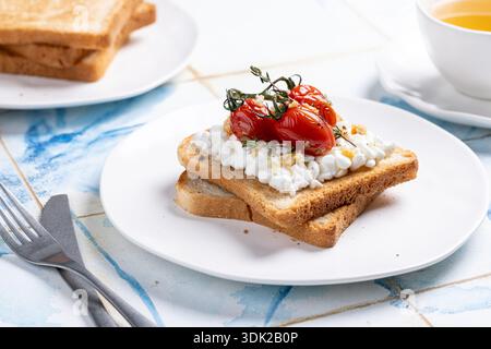 Eine Nahaufnahme eines leichten Frühstücks: Zwei Scheiben geröstetes Brot mit Hüttenkäse, garniert mit gerösteten Kirschtomaten auf der Weinrebe und Zweigen von Ihnen Stockfoto